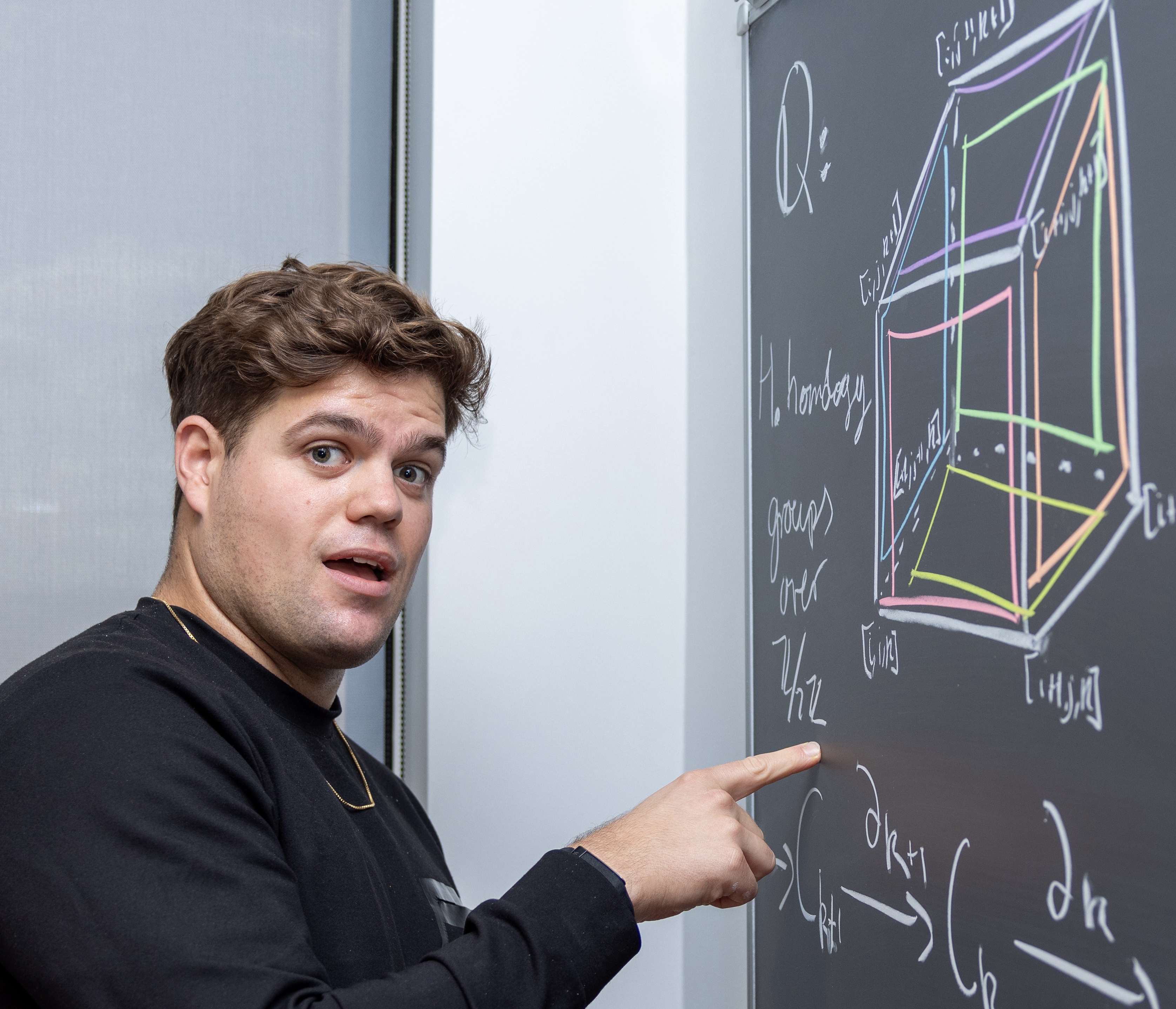 Yossi Bokor Bleile smiling and pointing at a blackboard with a cubical complex on it. Photo by Glenn Herping.